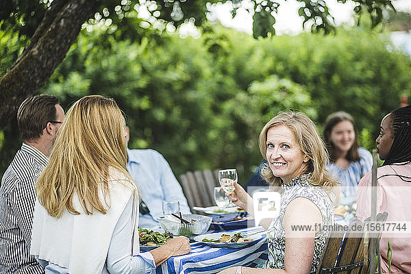 Lächelnde Frau hält ein Weinglas in der Hand  während sie mit Freunden auf einer Gartenparty sitzt