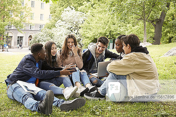 Lächelnder Teenager zeigt Freunden sein Smartphone  während er im Park sitzt