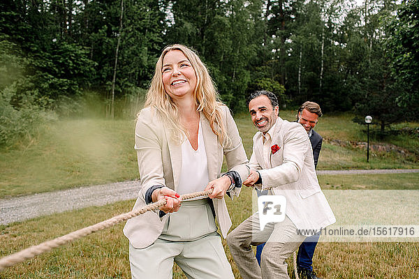 Smiling male and female colleagues pulling rope while playing tug-of-war in lawn