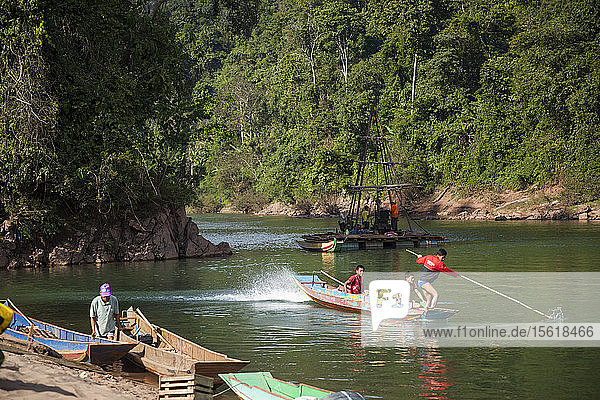Young men speed off in a motorboat past a drill rig collecting core samples on the Nam Ou River at the planned site for Dam #7 within Phou Den Din National Protected Area  Laos.