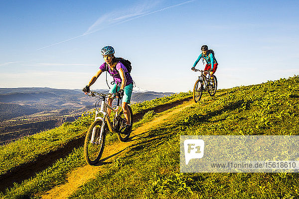 Two young women ride mountain bikes downhill on a single-track trail through green grass in early morning sunlight.