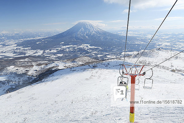 Vor dem Hintergrund des Vulkans Yotei sitzen eine Skifahrerin und ein Skifahrer in einem Sessellift  der sie zum Gipfel des Annupuri im Skigebiet Niseko United auf der japanischen Insel Hokkaido bringt. Niseko United besteht aus vier Skigebieten auf dem Annupuri (1.308 m). Der 100 km südlich von Sapporo gelegene Niseko Annupuri ist Teil des Quasi-Nationalparks Niseko-Shakotan-Otaru Kaigan und ist der östlichste Park der Niseko-Vulkangruppe. Hokkaido  die nördliche Insel Japans  liegt geografisch ideal in der Bahn der beständigen Wettersysteme  die die kalte Luft aus Sibirien über das Japanische Meer bringen. Dies führt dazu  dass viele der Skigebiete mit Pulverschnee überhäuft werden  der für seine unglaubliche Trockenheit bekannt ist. In einigen der Skigebiete Hokkaidos fallen durchschnittlich 14-18 Meter Schnee pro Jahr. Niseko ist die Pulverschneehauptstadt der Welt und als solche das beliebteste internationale Skigebiet in Japan. Es bietet ein unvergessliches Erlebnis für Skifahrer und Snowboarder aller Niveaus. Der Berg Yotei im Hintergrund wird oft auch als der Berg Fuji von Hokkaido bezeichnet.