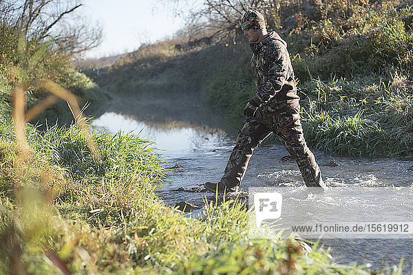 Junger erwachsener männlicher Jäger  der auf der Suche nach Rehen durch einen Bach läuft