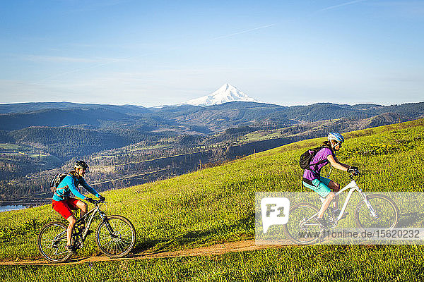 Two young women ride mountain bikes up a single-track trail with mountains and a snow-capped volcano in distance.