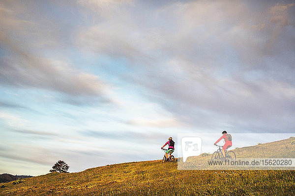 Two young women ride mountain bikes on a single-track trail through grassy meadow and early morning sky.
