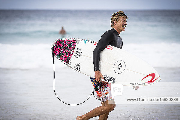 Side view of single smiling male surfer carrying surfboard along beach