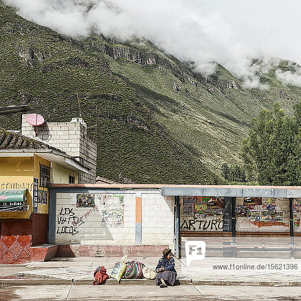 Eine peruanische Frau  die auf einen Bus wartet  mit einem sich im Hintergrund erhebenden Berg