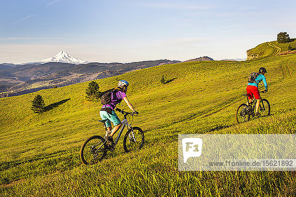 Two young women ride mountain bikes up a single-track trail through an open meadow with volcano in distance.