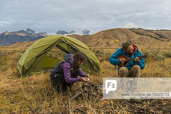 Eine Camperin bereitet das Frühstück auf einem Campingkocher zu  während ein Camper vor seinem Zelt sitzt und Mandoline spielt  Jackson  Wyoming  USA