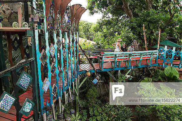 Besucher spazieren über eine farbenfrohe Brücke im Jade Seahorse - eine eklektische  künstlerische Anlage mit Zimmern zur Miete  einer Bar in einem Baumhaus  verrückten Strukturen  Wegen und Brücken auf der Insel Utila  Honduras.