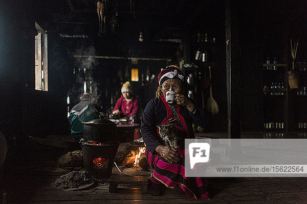 Porträt einer älteren Frau  die hinter einem Lagerfeuer sitzt  eine Katze auf dem Schoß hat und aus einer Tasse trinkt  Myanmar  Shan  Myanmar