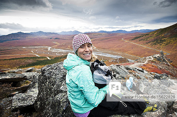 A girl in adventure gear sits on top of a mountain overloooking Denali National Park.