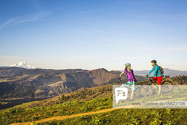Zwei junge Frauen machen auf ihren Mountainbikes eine Pause  während sie auf einem einspurigen Weg durch eine offene Wiese mit Fluss und Vulkan in der Ferne fahren.