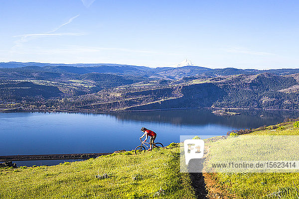 A man rides a mountain bike downhill on a single-track trail through green grass with a large river in the distance.
