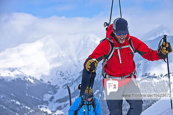 Ein Skifahrer und ein Snowboarder wandern auf einem Berg mit schneebedeckten Gipfeln in der Ferne.