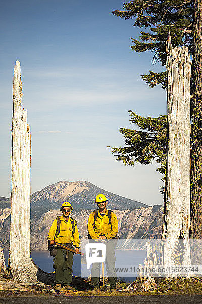 Zwei Feuerwehrleute in gelben Hemden stehen mit ihrer Ausrüstung zwischen zwei abgestorbenen Bäumen  im Hintergrund ein Bergsee. Crater Lake  Oregon  USA