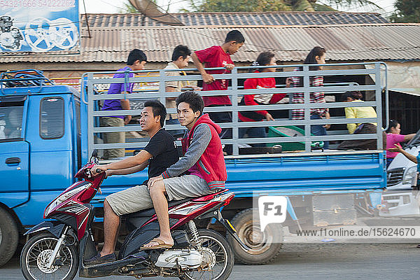 Junge Männer fahren gemeinsam auf einem Motorroller an einem Lastwagen vorbei  der auf einer belebten Straße in Luang Prabang  Laos  entladen wird.