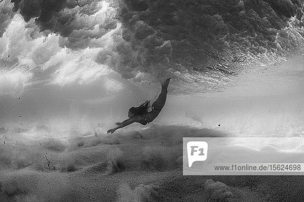 Schwarz-Weiß-Foto eines Mädchens im Bikini  das unter Wasser im Ozean schwimmt. Sie geht unter den Wellen über einen sandigen Boden  der von klarem Wasser bedeckt ist.