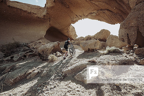 Mann geht in der Wüste mit seinem Mountainbike in Richtung eines Felsbogens  Teneriffa  Kanarische Inseln  Spanien