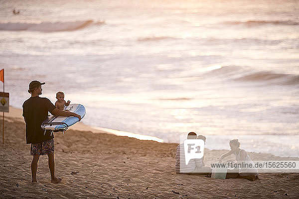 Surfer mit Baby auf Surfbrett und andere Menschen am Strand bei Sonnenuntergang  Oahu  Hawaii-Inseln  USA