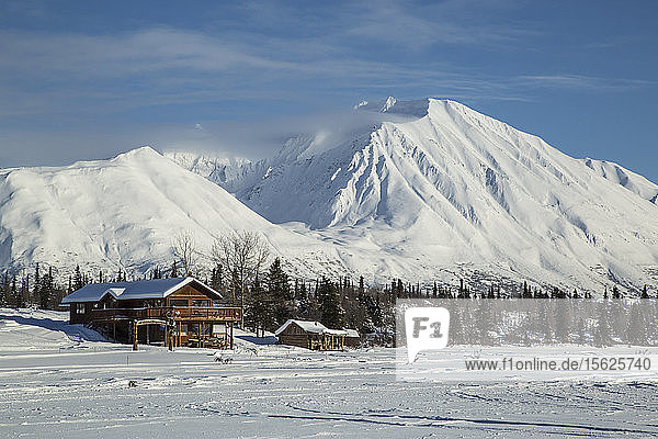 Log cabins in Alaska sit at the base of a frozen lake with snowy mountains in the background.