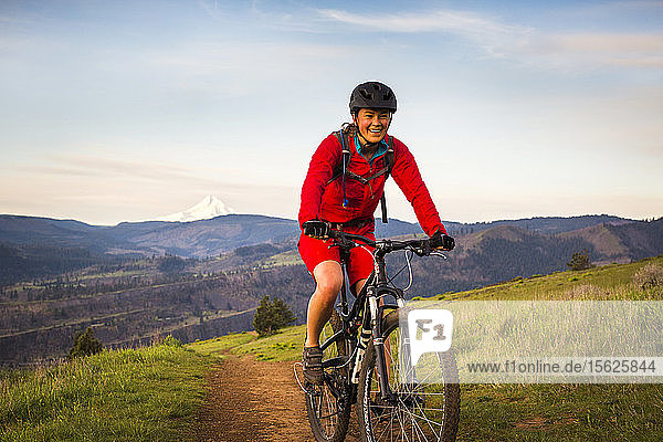 A young woman in a red coat rides a mountain bikes on single-track trail through green grass in early morning light.
