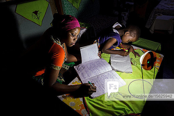 Mforo  Tansania ein Dorf in der Nähe von Moshi  Tansania. Mit einer Solar Sister-Solarlaterne lernen mehrere Kinder der Solar Sister-Unternehmerin Fatma Mziray?????? nachts. Ihre ältere Tochter Zainabu Ramadhani (19 Jahre) ist links und die mittlere Tochter Sabrina Ramadhani (8 Jahre) ist rechts. Fatma Mziray ist eine Solar Sister-Unternehmerin  die sowohl saubere Kochherde als auch Solarlaternen verkauft. Fatma hörte von einem Entwicklungshelfer von Solar Sister von den Kochern und beschloss  einen auszuprobieren. Der Rauch vom Kochen auf ihrem traditionellen Holzherd mit Brennholz verursachte ihr viele gesundheitliche Probleme  ihre Lungen waren verstopft  ihre Augen brannten und ihr Arzt sagte ihr  dass sie nicht mehr auf diese Weise kochen dürfe. An manchen Tagen fühlte sie sich so schlecht  dass sie nicht zum Kochen gehen konnte. Fatma sagte: ??????Um für eine Familie zu kochen  Frühstück  Mittag- und Abendessen zuzubereiten  musste ich früher jeden Tag eine große Ladung Holz sammeln. Jetzt  mit dem neuen Herd  reicht die gleiche Ladung Holz für bis zu drei Wochen Kochen. ??????Durch die zusätzliche Zeit kann ich mein Geschäft ausbauen. Außerdem habe ich mehr Zeit für meine Familie. Ich kann das Studium meiner Kinder ?????? überwachen. All das trägt zu einer glücklicheren Familie und einer besseren Beziehung zu meinem Mann bei. Seitdem ich den sauberen Herd benutze  ist niemand mehr krank geworden oder musste wegen einer Grippe ins Krankenhaus. ?????? Fatma sieht sich selbst als Helferin ihrer Gemeinschaft  denn sie sieht nicht mehr  dass die Menschen  denen sie Kochherde verkauft hat  rote Augen haben  husten oder krank sind  wie es früher der Fall war. Sie konnte das Schulgeld für ihre Kinder bezahlen  Dinge für den Haushalt und eine Kuh kaufen. ??????Was mich dazu bringt  jeden Morgen früh aufzustehen  meine Kocher zu nehmen und in mein Geschäft zu gehen  ist die Möglichkeit  meine Familie zur Schule zu bringen sowie Lebensmittel und andere Familienbedürfnisse zu besorgen.??????