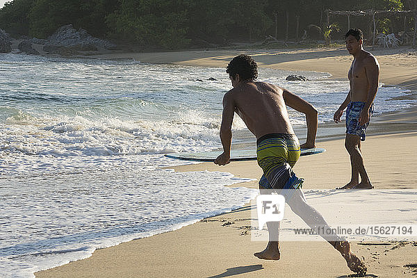 Ein junger männlicher Skimboarder rennt auf das Meer zu  während ein anderer Strandbesucher in Mazunte  Oaxaca  Mexiko  zusieht.