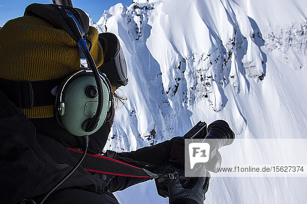 Ein Snowboarder-Filmer hängt an einem sonnigen Tag in Haines  Alaska  seitlich aus einem Hubschrauber  um Snowboarder zu filmen  die ein Gesicht schneiden.