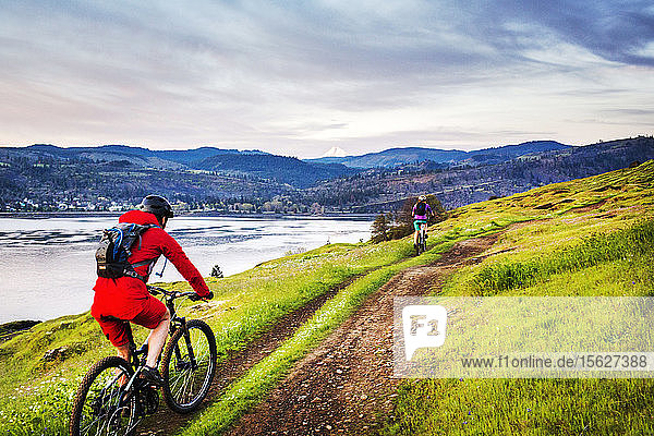 A young woman in red coat rides a mountain bike on single-track trail through green grass with a large river and mountains in distance.