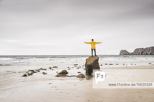 Junger Mann in gelber Regenjacke am Strand und auf Felsen stehend Bretagne Frankreich