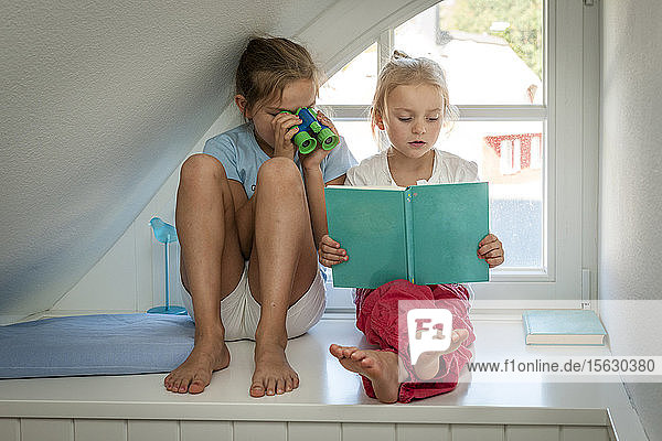Girl with toy binoculars looking at sister with book at the window