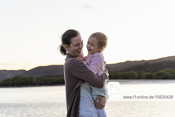 Glückliche Mutter trägt ihre Tochter bei Sonnenuntergang auf einem Steg