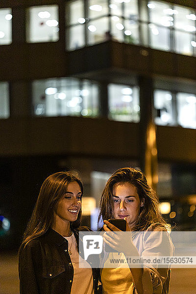 Zwei Freunde benutzen das Smartphone bei Nacht  mit den Lichtern der Stadt im Hintergrund