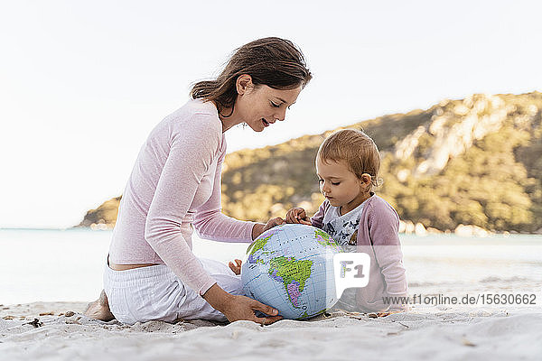 Frau und kleine Tochter sitzen am Strand und schauen gemeinsam auf den Strandball der Erde