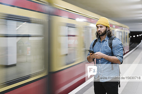 Portrait of man listening music with earphones and smartphone while waiting at platform  Berlin  Germany