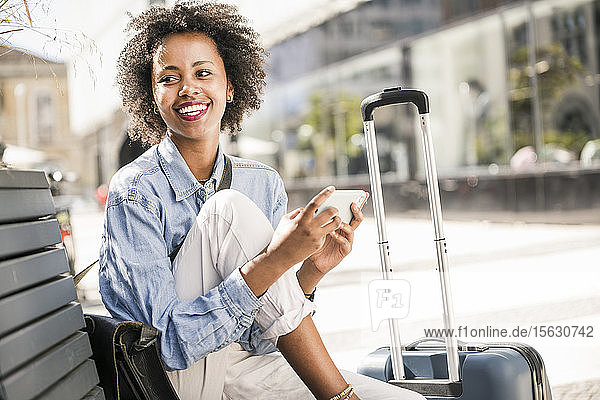 Glückliche junge Frau sitzt auf einer Bank mit Handy und Koffer