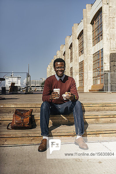 Young man sitting on stairs in the city  eating hamburger  drinking coffee