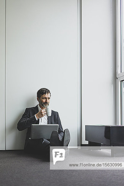 Reifer Geschäftsmann im Büro  der mit Kaffeetasse und Laptop auf dem Boden sitzt