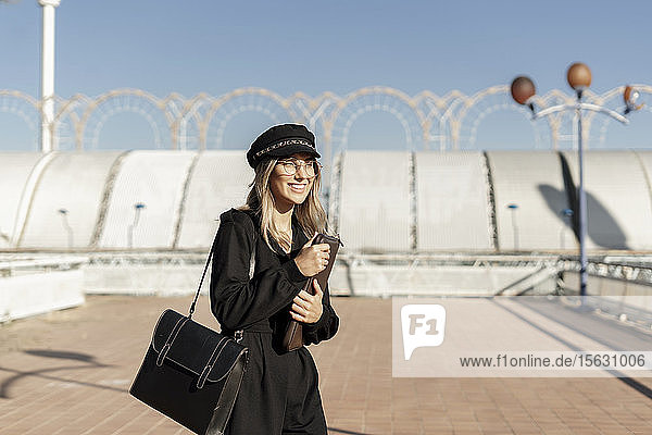 Junge blonde Geschäftsfrau mit schwarzer Matrosenmütze und Laptoptasche in der Hand