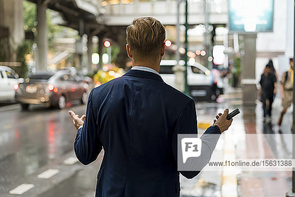 Junger Geschäftsmann hält Smartphone in Bangkok an einem regnerischen Tag