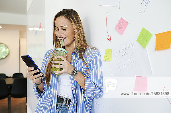 Fröhliche lockere junge Geschäftsfrau mit Getränk und Handy in einem Büro
