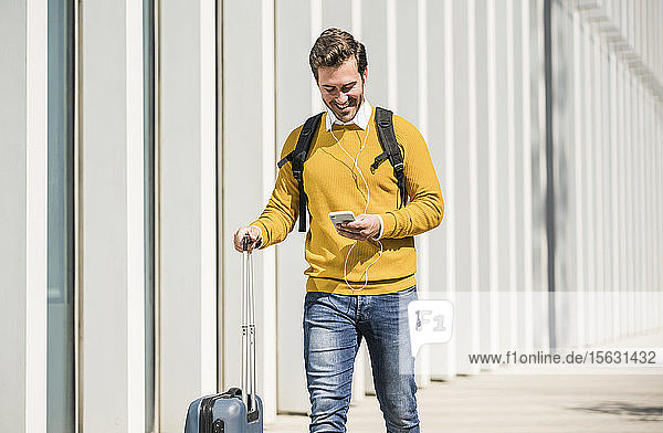 Lächelnder junger Mann mit Rollkoffer und Kopfhörern beim Telefonieren mit dem Handy in der Stadt
