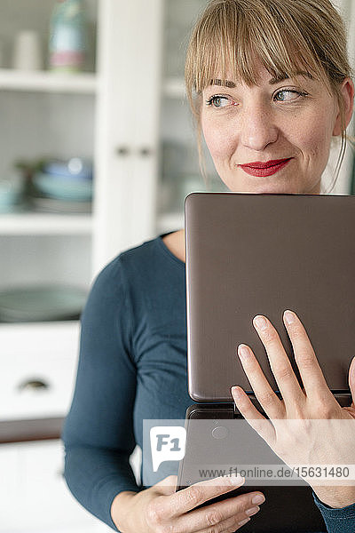 Porträt einer lächelnden Frau mit Laptop in der Küche  die in die Ferne schaut