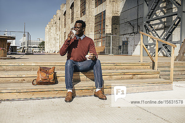 Junger Mann sitzt auf einer Treppe in der Stadt  isst Hamburger  trinkt Kaffee