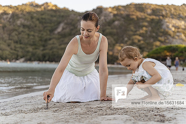 Mutter und Tochter zeichnen mit kleinen Stöckchen im Sand am Strand