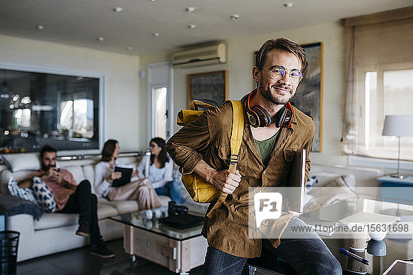 Portrait of smiling man with backpack  headphones and laptop with friends in background