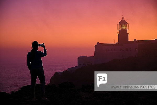 Portugal  Algarve  Silhouette einer Person  die in der Morgendämmerung per Smartphone Fotos vom Leuchtturm von Cape Saint Vincent macht