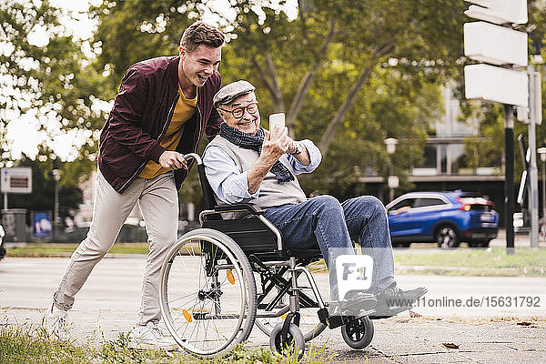 Lächelnder junger Mann schiebt glücklichen älteren Mann mit Smartphone im Rollstuhl