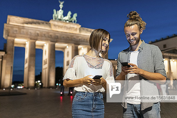 Glückliches Paar benutzt Smartphones am Brandenburger Tor bei Nacht  Berlin  Deutschland